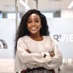 A confident woman with crossed arms standing in an office environment, with the word "interview" visible in the background.