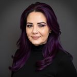 A person with long purple hair and purple eye makeup wearing a black top against a dark background.