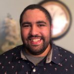 A man with a beard and mustache smiles at the camera, wearing a dark shirt with a light pattern. A blurred clock is visible in the background.