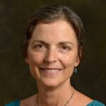 A woman with short hair smiles, wearing a teal top, blue earrings, and a delicate necklace. The background is a plain, dark color.