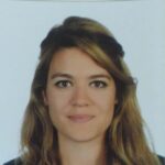 A woman with long, light brown hair and a black top is facing forward against a plain white background.
