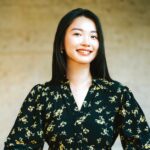A woman with long dark hair, wearing a black blouse with a yellow floral pattern, stands smiling against a plain beige background.