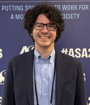 A person with curly dark hair and glasses smiles at an event, standing in front of a blue backdrop with white text and “#ASA26” displayed.