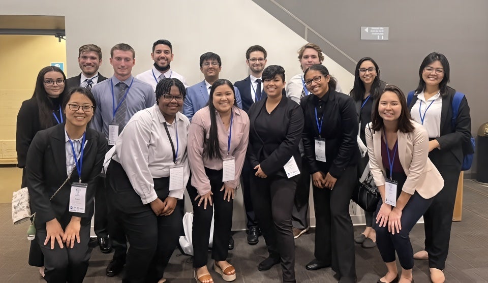 A group of fifteen professionally dressed young adults pose together indoors, wearing name badges and smiling at the camera.