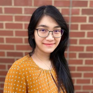 A woman with long dark hair and glasses smiles, wearing a yellow patterned blouse, standing in front of a brick wall.