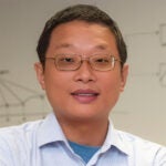 A person wearing glasses and a light blue collared shirt, standing in front of a blurred background with diagrams.