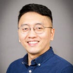 A man with short black hair, glasses, and a navy blue button-up shirt smiles at the camera against a plain gray background.