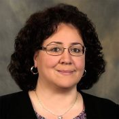 A woman with curly dark hair, wearing glasses, hoop earrings, and a necklace, is pictured against a neutral, mottled background. She is dressed in a dark top with a patterned neckline.