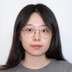 Woman with long hair and glasses wearing a gray shirt against a plain background.