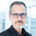 Man with short gray hair, a goatee, and blue glasses wearing a black blazer standing indoors in front of large bright windows.