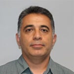 A middle-aged man with short dark and gray hair is wearing a green plaid button-up shirt and looking straight at the camera against a plain light background.