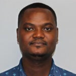 A man with short hair and a trimmed beard, wearing a blue collared shirt with a white pattern, facing forward against a plain background.