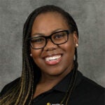 Smiling woman with long braids and glasses wearing a black collared shirt poses in front of a gray background.