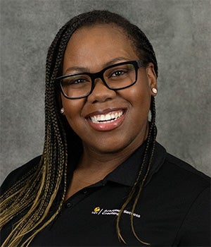 Smiling woman with long braids and glasses wearing a black collared shirt poses in front of a gray background.