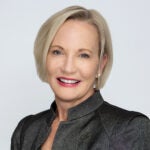 A woman with short blonde hair, wearing a dark blazer, smiling against a plain light background.