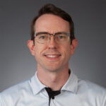 A man wearing glasses and a white collared shirt with a UCF logo stands in front of a plain gray background, looking at the camera and smiling slightly.