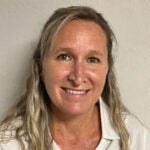 A woman with long, wavy blonde hair, wearing a white collared shirt, smiles at the camera against a plain light-colored background.