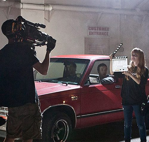 Film crew shooting a scene with actors in a car; cameraman capturing footage while a crew member holds a clapperboard.