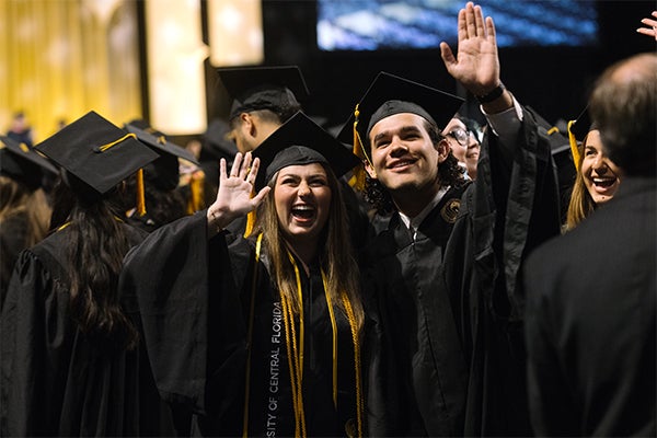 Two graduates in caps and gowns smile and wave during a graduation ceremony, surrounded by other graduates.