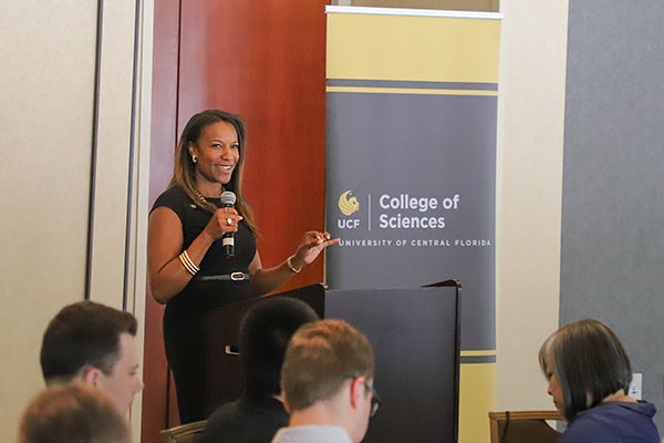 A woman speaks at a podium with a microphone beside a sign that reads "UCF College of Sciences University of Central Florida" as attendees listen.