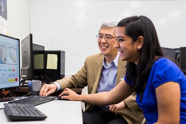 Two people sit at a desk, looking at a computer screen and smiling, with office equipment and whiteboard in the background.