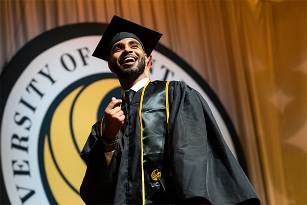 A graduate in cap and gown smiles on stage in front of a University of Central Florida emblem during a commencement ceremony.