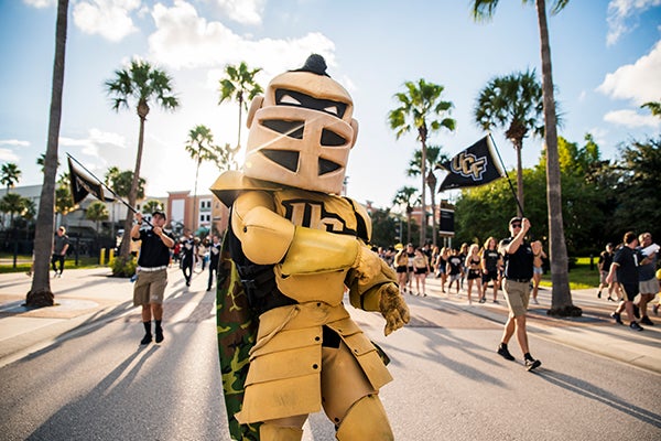 A person in a knight mascot costume leads a parade with people holding UCF flags and palm trees lining the street in the background.