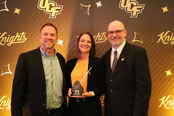 Three adults in business attire stand together smiling; the woman in the center holds an award. The background displays "UCF" and "Knights" logos.