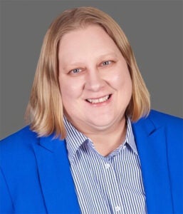 A person with shoulder-length blond hair, wearing a blue blazer over a striped shirt, smiling at the camera against a plain gray background.