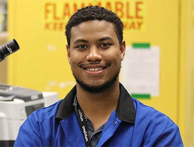 A person wearing a blue lab coat and black lanyard stands smiling in front of a yellow cabinet labeled "Flammable Keep Away." A microscope is visible to the left.