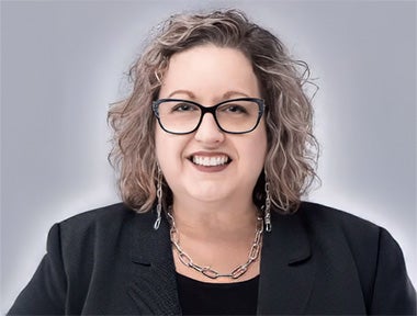 A woman with curly, shoulder-length hair and glasses smiles at the camera. She is wearing a black jacket, a black top, a necklace, and earrings, against a light grey background.