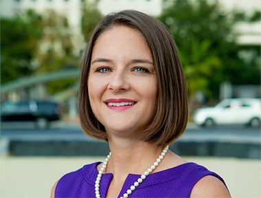 A woman with short brown hair wearing a purple dress and a pearl necklace stands outdoors, smiling at the camera. Cars and trees are visible in the blurred background.