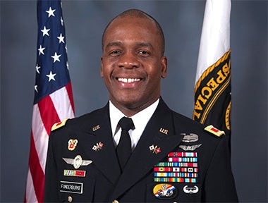 A US Army officer in full dress uniform stands in front of the American flag and a military flag, smiling at the camera.