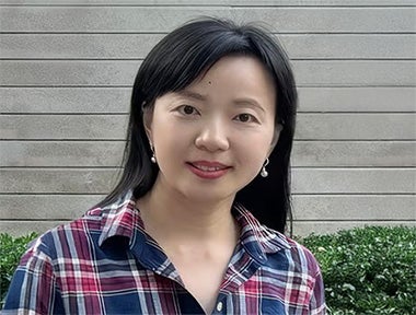 A woman with straight black hair, wearing a plaid shirt and pearl earrings, stands in front of a hedge and a horizontal wooden fence.