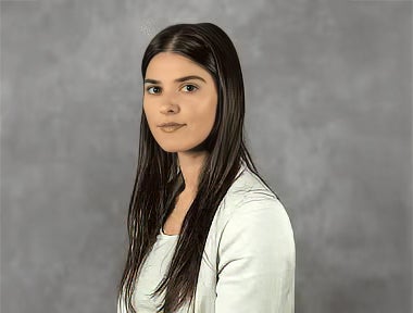 A woman with long straight brown hair wearing a light-colored blazer poses in front of a gray background.