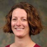 A woman with short, wavy brown hair and a maroon top smiles at the camera against a mottled green background.