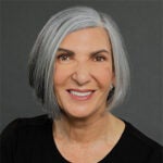 A woman with straight gray hair in a bob cut, wearing a black top, smiles at the camera against a plain dark background.