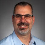A middle-aged man with short dark hair, glasses, and a beard, smiling, wearing a light blue collared shirt against a plain gray background.