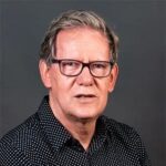 An older man with short gray hair and glasses wearing a black shirt with white dots, posing in front of a plain dark background.