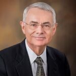 An older man with gray hair and glasses wearing a dark suit, white shirt, and patterned tie, poses against a brown gradient background.
