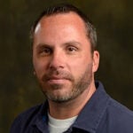 A man with short hair and a trimmed beard wearing a dark collared shirt and white t-shirt poses against a dark, neutral background.