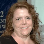 A woman with curly brown hair smiling at the camera, standing indoors in front of a blue and white background.