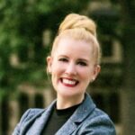 A woman with blonde hair in a bun, wearing a gray blazer and black top, smiles while standing outdoors with blurred greenery in the background.