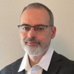 A man with short hair, glasses, and a beard wearing a dark suit jacket and white collared shirt, standing against a plain light-colored wall.