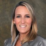 Woman with straight blonde hair wearing a grey blazer, smiling at the camera against a neutral, textured background.