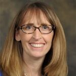 A woman with straight, shoulder-length light brown hair, wearing glasses, a blue top, and a silver necklace, smiles in front of a plain, blurred background.