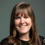 A woman with straight brown hair and bangs, wearing a black top with pearl details on the sleeves, smiles at the camera against a plain background.