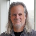 A man with long gray hair and a gray beard wears a gray UCF Lou Frey Institute shirt and stands indoors in front of a window.