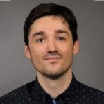 A man with short dark hair and a trimmed beard wearing a dark button-up shirt with a subtle pattern poses against a plain gray background.
