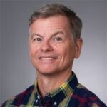A middle-aged man with short gray hair wearing a plaid shirt, smiling at the camera against a plain gray background.